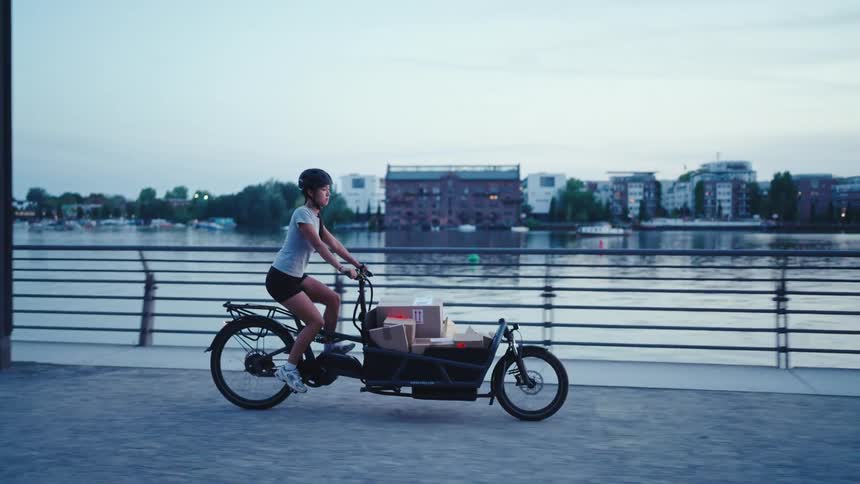 A young woman pedaling a long cargo bike loaded with packages along a riverway path