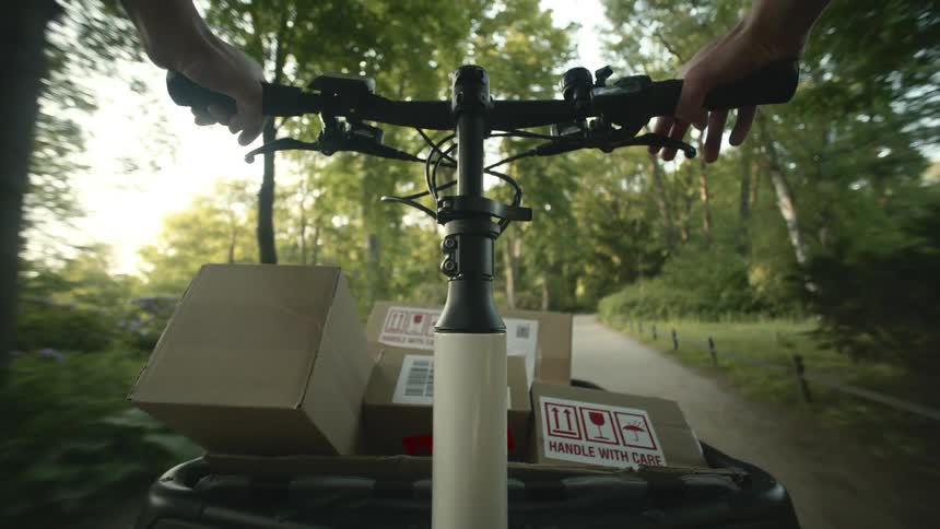 POV: A person riding a bike with a cargo carrier full of boxes labeled handle with care on a sunny day in a park