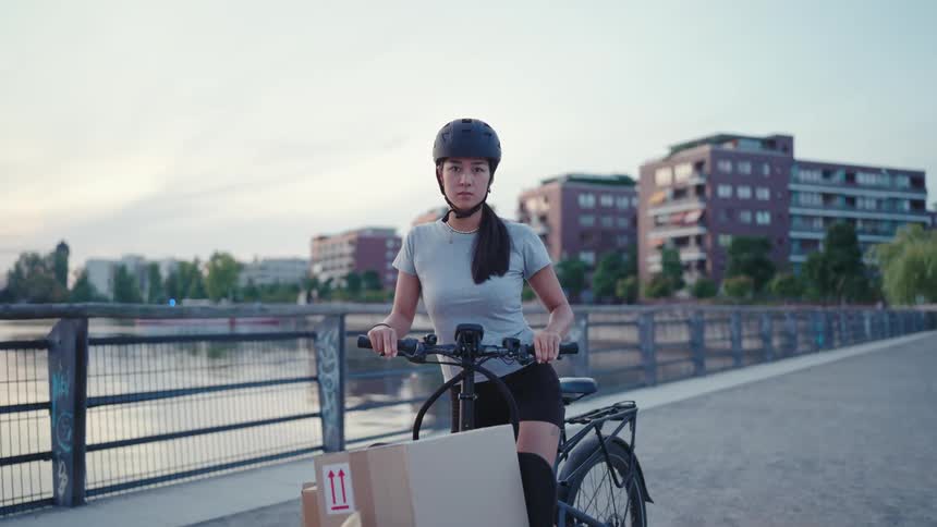 Young woman on an electric bike with a load of boxes standing on a bridge by a lake