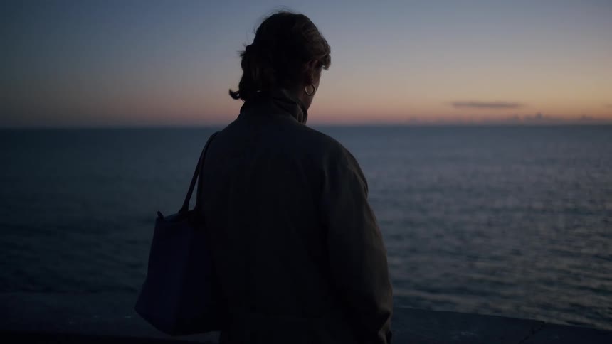 Woman with a pony tail a bag on her shoulder and a coat is silhouetted against the sky and vast ocean at sunset