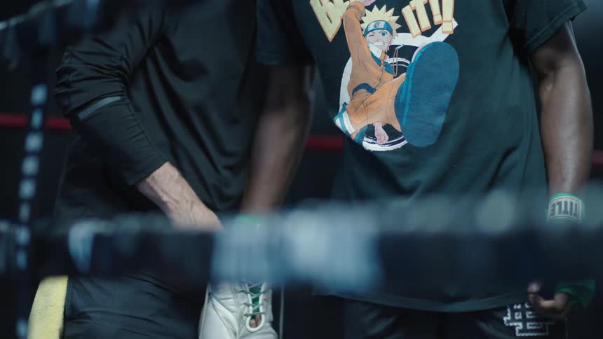 Man helps boxer put on his gloves while they stand in a boxing ring