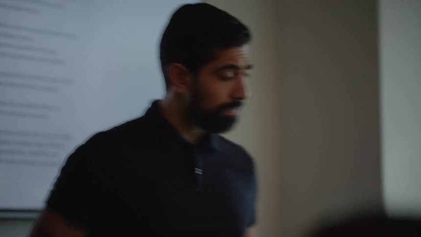 Coworker sitting at desk talks to a bearded man presenting to a small group in a meeting room with a large screen displaying a document behind him