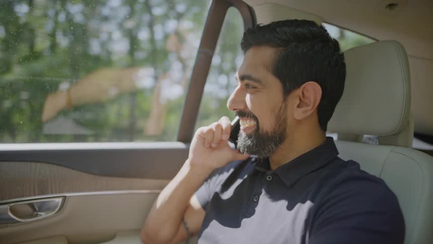 An indian man smiles as he talks on a smartphone while riding in a car through a suburb