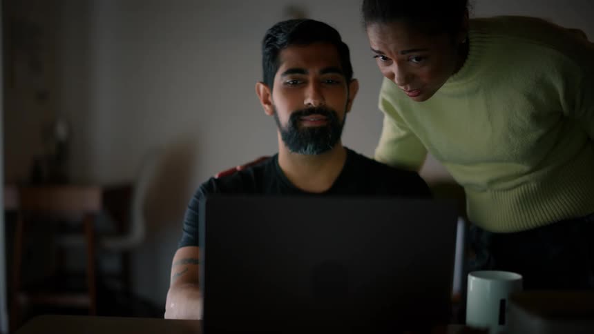 A man smiles while seated at his laptop and talking to his spouse