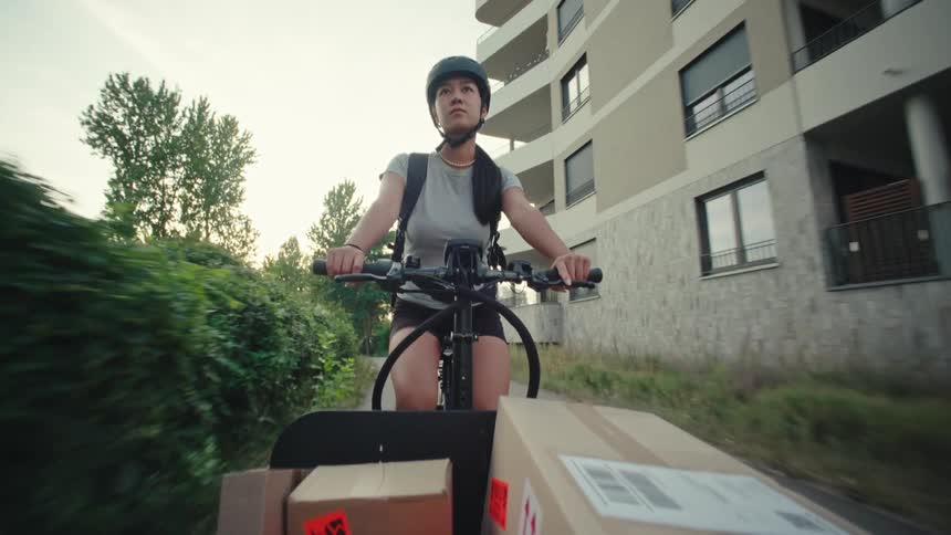 Woman pedals a cargo bicycle holding packages past apartments while delivering packages POV