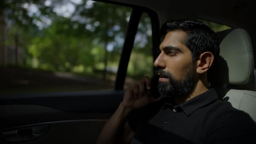 Man having a phone call sits in a car driving past a building near a grove