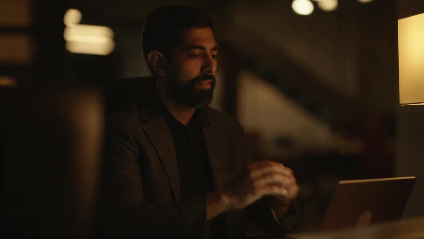 A bearded man in a brown blazer sits at his desk working on his laptop late into the night