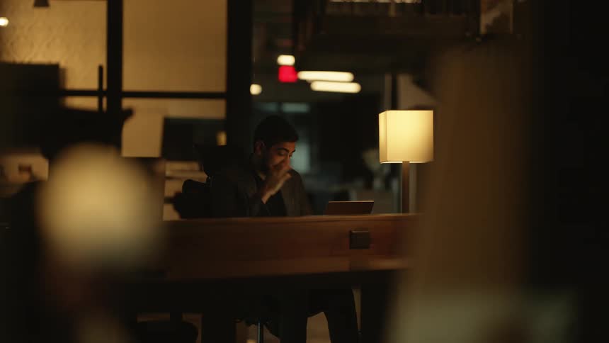 Male worker rubs his hand over his face while he works late on his laptop in a dark office