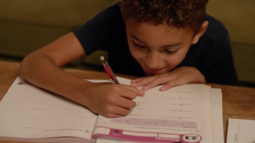 A young boy smiles as he sits in front of a coffee table and does homework from school