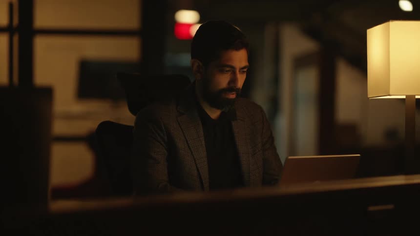 A man with a beard works on his laptop in a dimly lit office