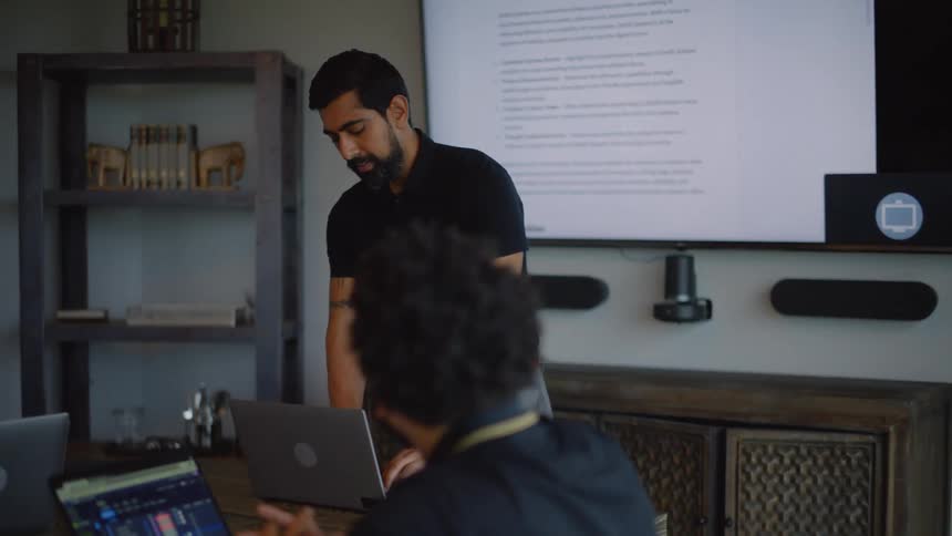 A bearded man wearing a black shirt and gray pants walks into the meeting room to present to his coworkers