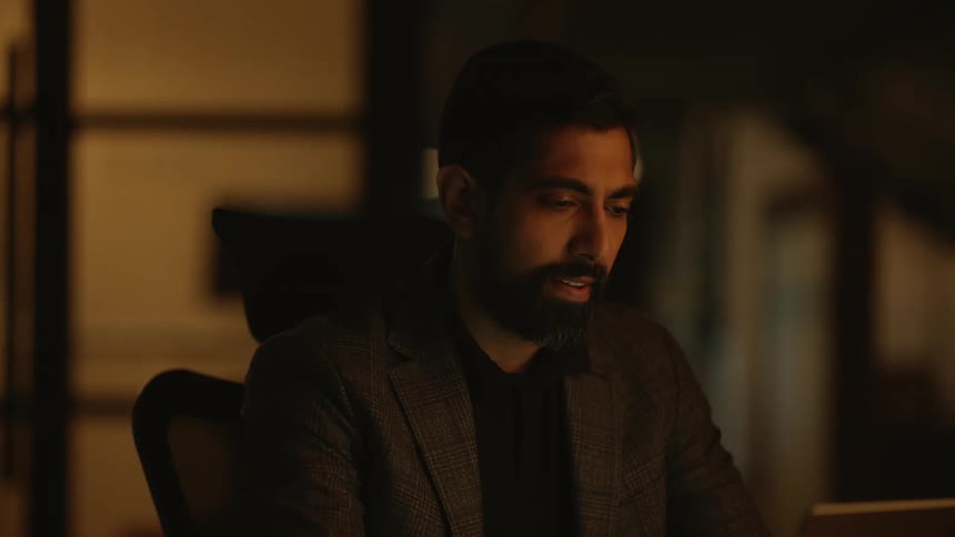 A man is looking at his laptop in a dark room at night in an office