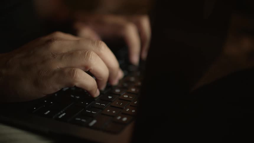 A person types on a laptop keyboard illuminated in the darkness