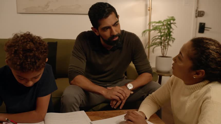 A father and mother talk while their son studies on the living room table