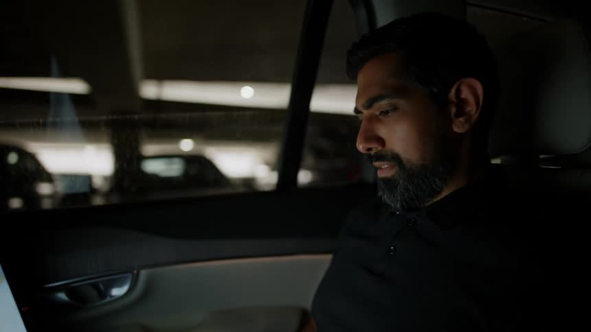 A bearded man uses his laptop while riding in a car in a parking garage