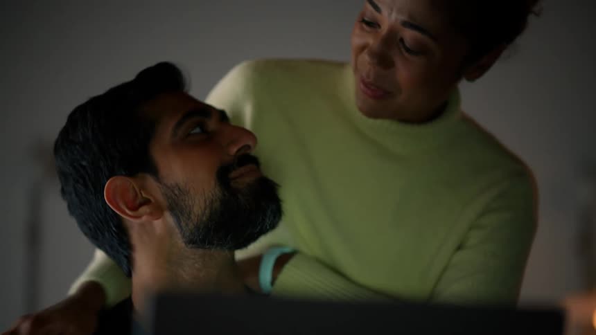 Woman hugs man sitting in front of a computer and talks to him