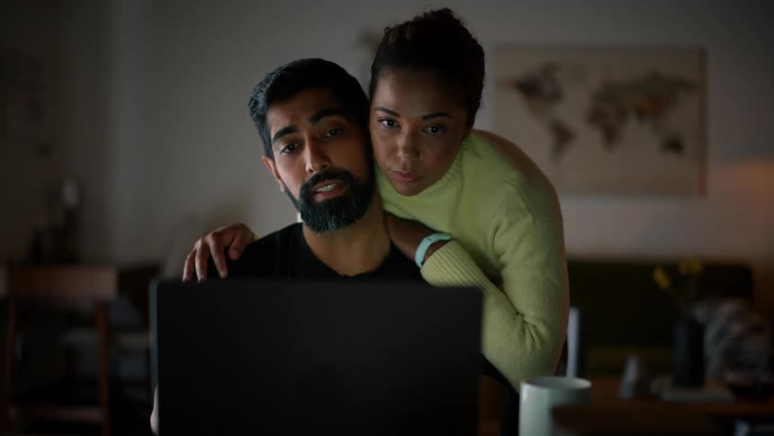 A man and a woman closely interacting with the woman leaning over his shoulder as they both focus intently on a laptop screen in a dimly lit room