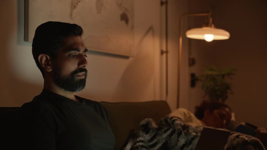 A man is sitting at home using his laptop at night while a woman lays next to him