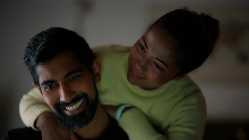 A woman leans on a mans shoulders as he types on a laptop and smiles