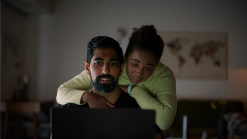 Woman wearing a green shirt cuddles bearded man in a black t shirt working on laptop at a desk