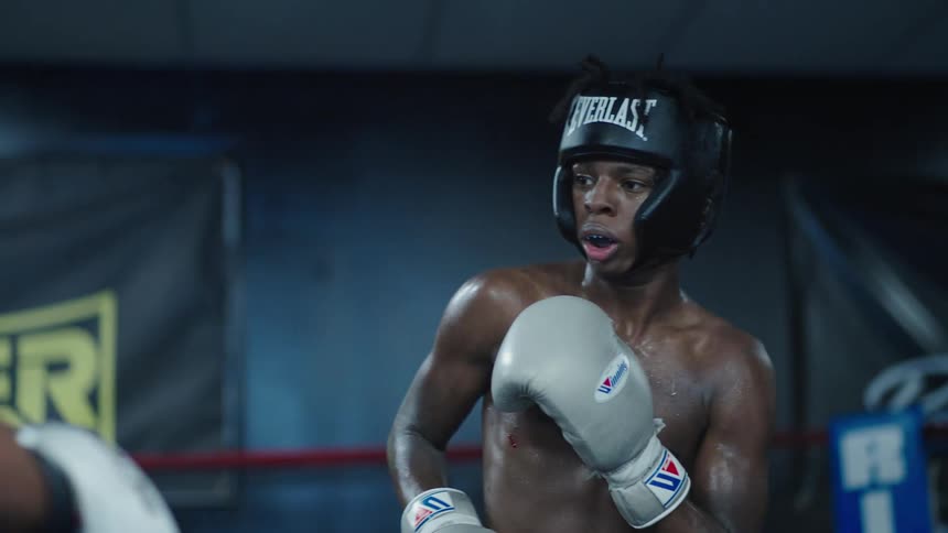 A young boxer wearing headgear and gloves facing an opponent in sparring session with a focused and determined expression