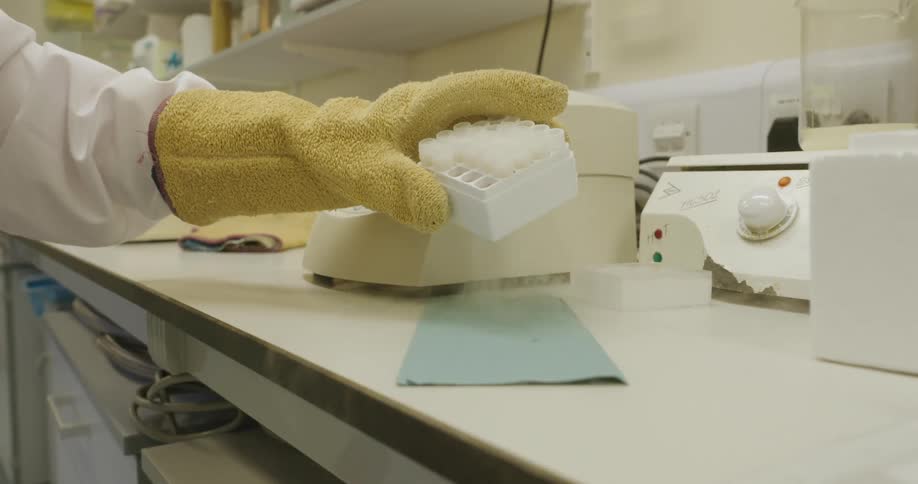 A person wearing a yellow protective glove places a white multiwell sample tray on a laboratory bench