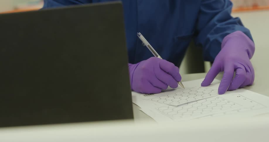 A person in a blue lab coat with purple gloves fills out a document using a pen