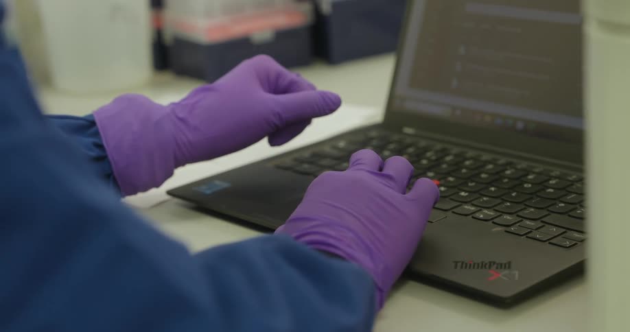 A pair of hands wearing purple gloves typing on a black thinkpad laptop