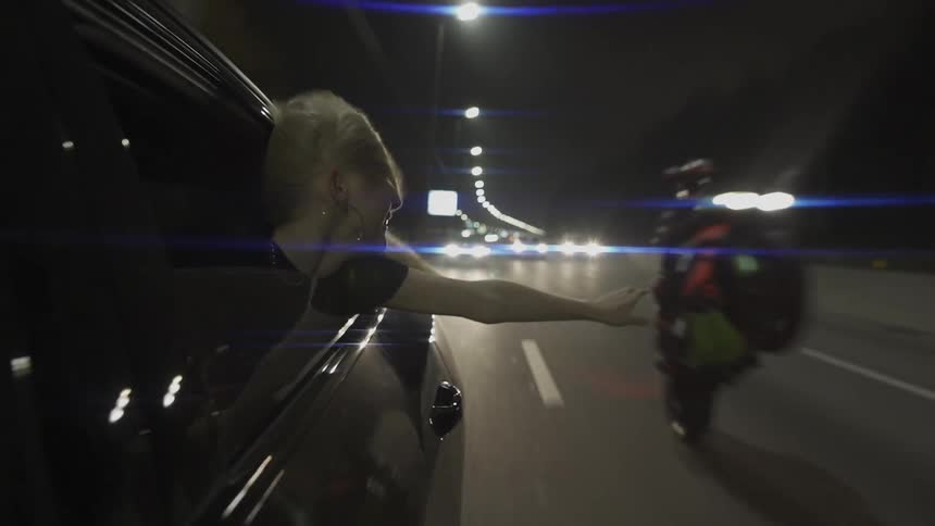 A young woman extends her arm out of a car window reaching for a motorcycle rider during nighttime
