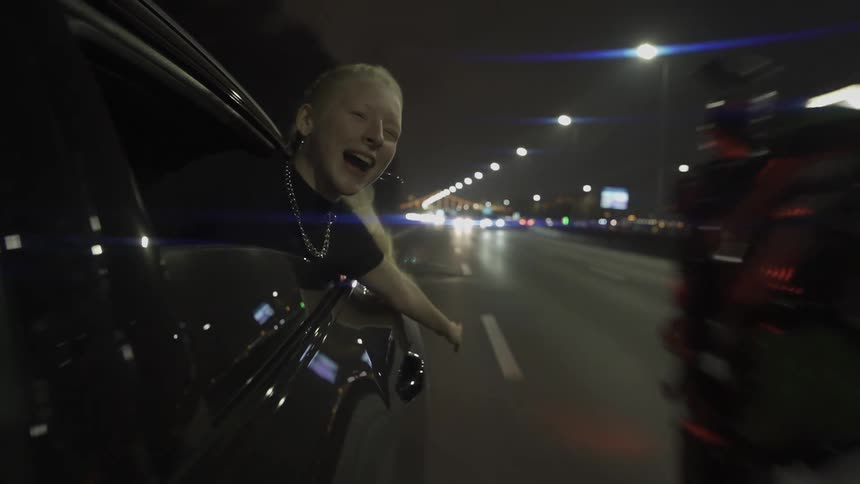 Woman in a car window with a motorcycle next to her doing a wheelie on the highway at night