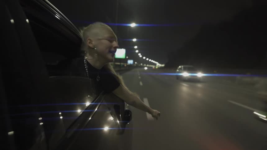 Blonde woman hanging out of a car window smiles as motorcyclist pops a wheelie while passing on the highway at night