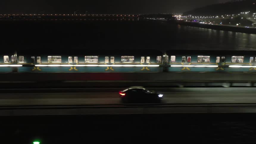 A night scene featuring a train with illuminated windows moving parallel to a road with a cars headlights on