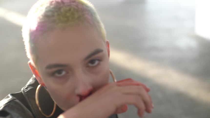 A young woman with hoop earrings and colored hair arises from a floor and wipes away blood from her nose