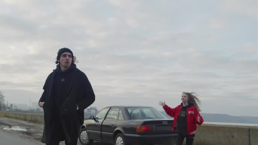 Man walks away from a car parked on a rural waterfront road as woman gestures angrily and follows