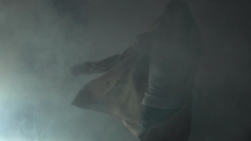 A woman wearing a trench coat swings her arms in a smoky room