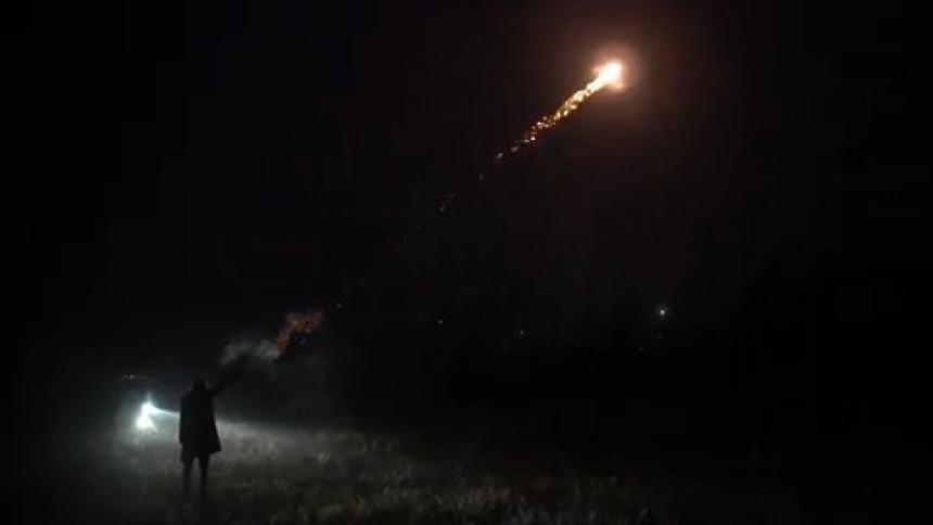 Person setting off fireworks in a field at night with a car in the background with its headlights on