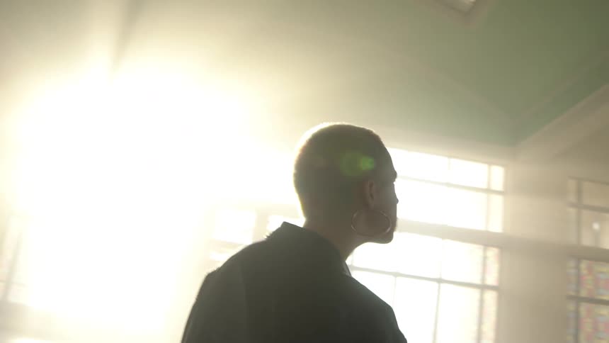 A young woman with short hair and a black coat looks around a large empty building as the sun shines through a window