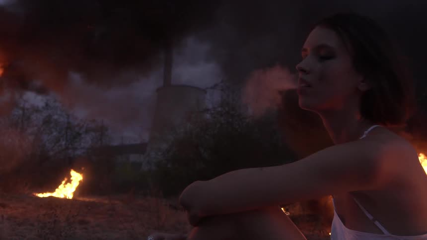 Fires burn around a woman smoking in a field near a power plant after dusk