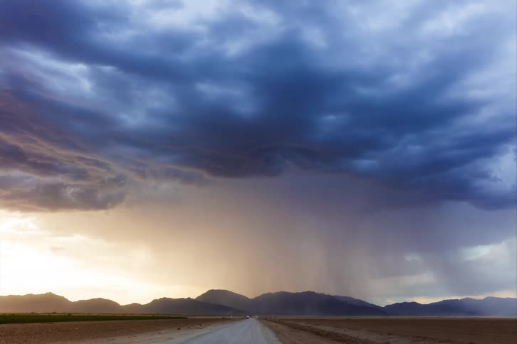 Dust flows across a rural roadwhile a storm front moves towards the nearby mountains