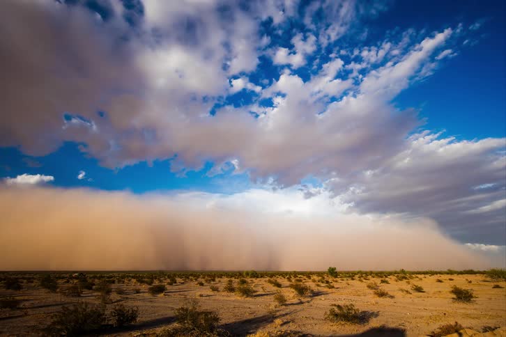A quick sandstorm passing through a desert on a day with blue sky and white clouds