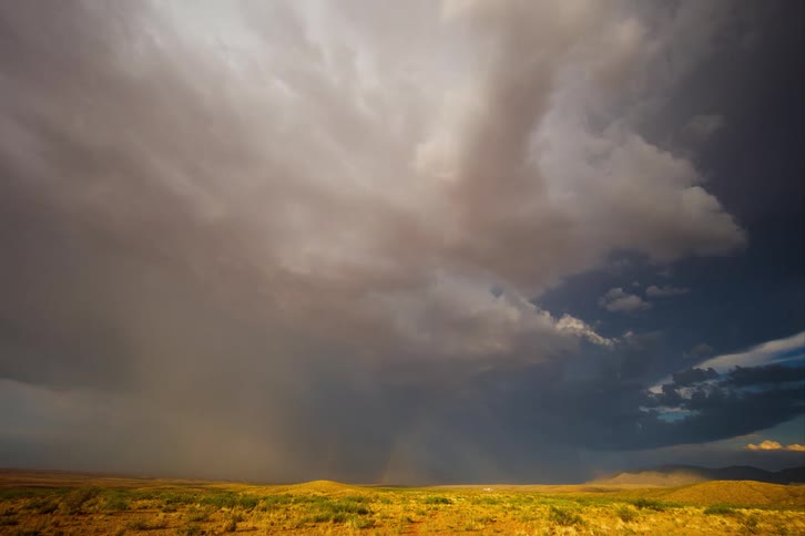 Rainbows appear against dark storm clouds over sunlit hills