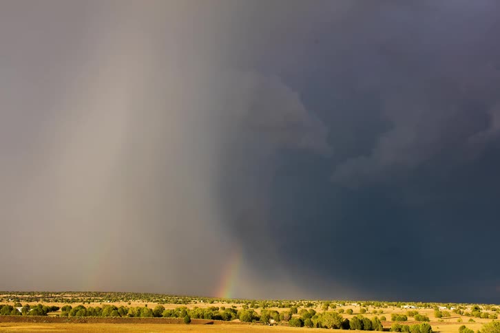Stormy sky with visible rain and a partial rainbow in the background in a field