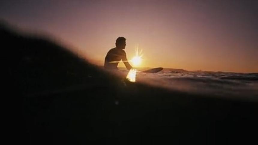 A surfer sits on his board silhouetted against a setting sun