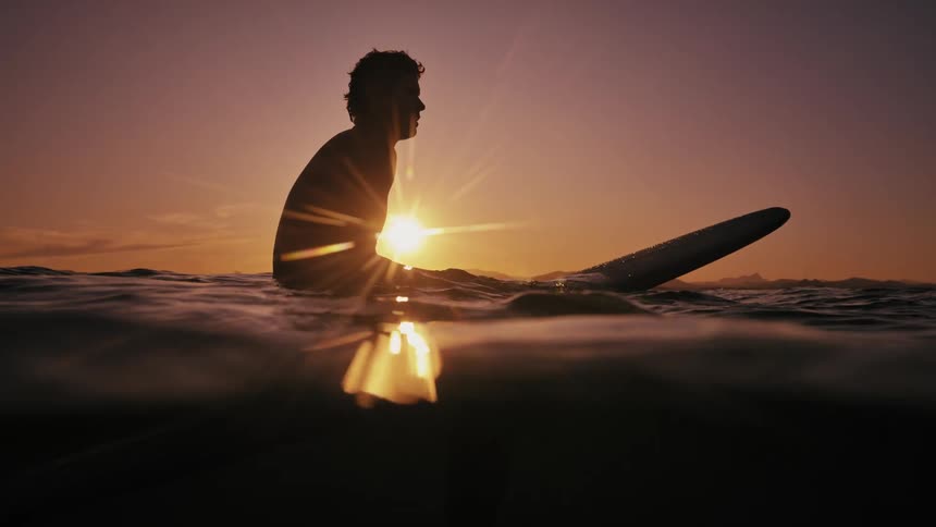 Silhouette of a man sitting and splashing water on his face while on his surfboard in the sea with the sun on the horizon