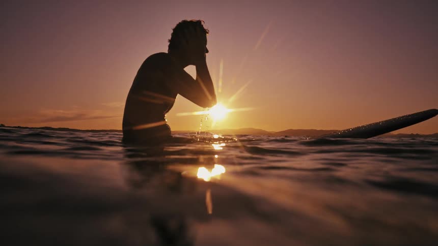 A silhouette of a man sitting on a surfboard splashing water on his face in the ocean as the sun sets