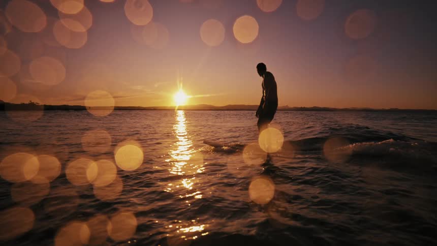 Male surfer standing on a surfboard is silhouetted by the sun sitting near the horizon in a red sky