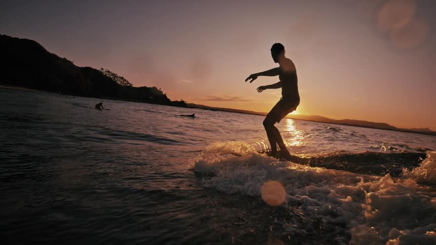 People in the sea with a man surfing on a board and the sunset on the horizon
