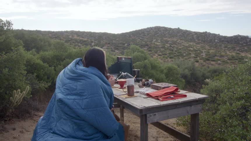 Couple sitting drinking coffee at a table while lying on a blue blanket amidst a wooded hill