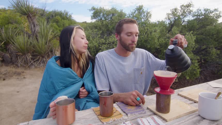 Man and woman sitting at a table on a hill surrounded by trees and preparing coffee