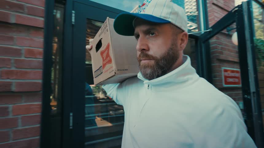 A man with a beard walks into a bar carrying a case of beer